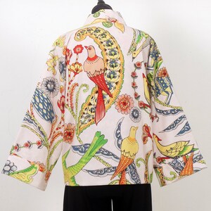 K&ouml;nnte beinhalten: Hellfarbene Jacke mit einem lebendigen Vogel- und Blumenmuster. Die Jacke hat ein farbenfrohes Design mit roten, gelben, gr&uuml;nen und blauen Akzenten. Die Jacke wird auf einer schwarzen Schaufensterpuppe pr&auml;sentiert.