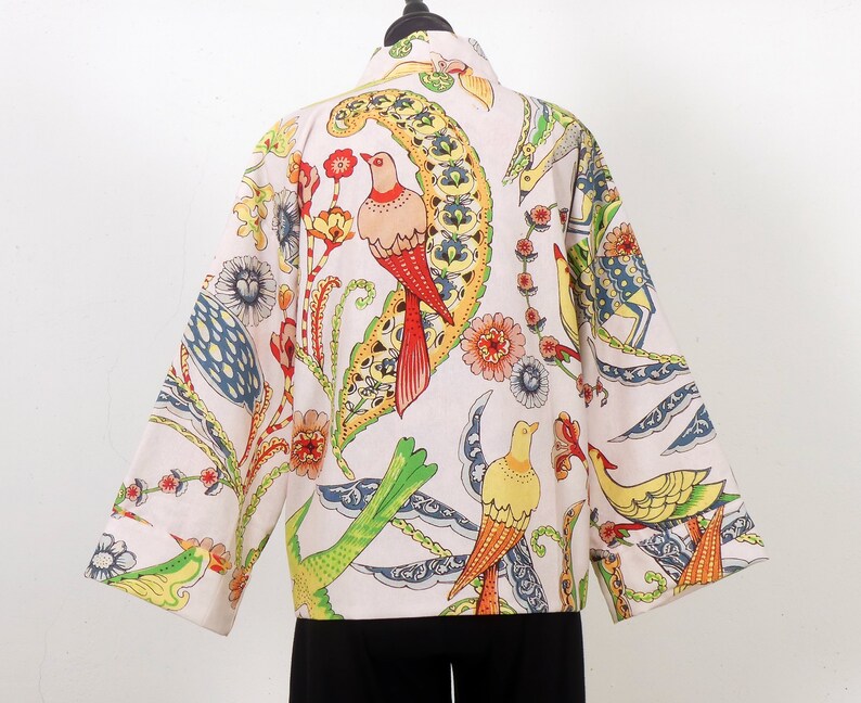 K&ouml;nnte beinhalten: Hellfarbene Jacke mit einem lebendigen Vogel- und Blumenmuster. Die Jacke hat ein farbenfrohes Design mit roten, gelben, gr&uuml;nen und blauen Akzenten. Die Jacke wird auf einer schwarzen Schaufensterpuppe pr&auml;sentiert.