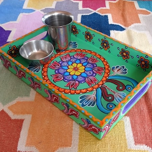 Peut inclure: Un plateau de service en bois peint à la main, avec une base verte, orné d'un motif floral central de mandala aux couleurs vives. Le plateau est de forme rectangulaire avec des poignées et comprend deux petits bols en acier inoxydable.