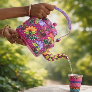 Puede incluir: Una tetera pintada a mano, vibrante, en tonos rosa, morado, naranja y verde, adornada con motivos florales. La tetera vierte agua en una taza a juego. El asa y el pico están intrincadamente diseñados, creando un juego de té único y artístico.