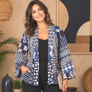 Peut inclure: Une femme porte une veste de style kimono à motifs bleus et blancs avec un haut noir et un pantalon noir. La veste présente un motif patchwork avec des motifs géométriques et abstraits. L'arrière-plan comprend une peinture et des objets décoratifs.