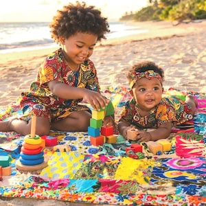 Peut inclure: Deux enfants sur une plage jouent avec des jouets en bois colorés. Ils portent des tenues assorties à motifs et sont assis sur une couverture patchwork vibrante. Un empileur d'anneaux en bois et des blocs de construction sont visibles.