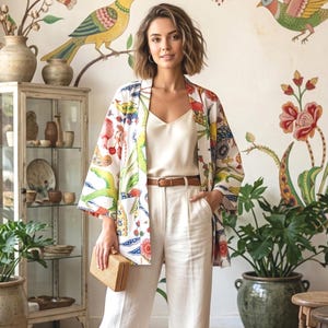 Peut inclure: Une femme porte un kimono blanc orné de motifs floraux et d'oiseaux colorés, un caraco blanc et un pantalon blanc. Elle tient une petite pochette tissée. L'arrière-plan présente une fresque florale et une vitrine.