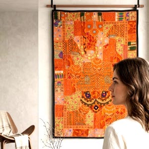 Peut inclure: Une tenture murale patchwork aux couleurs vives, orange et jaune, avec des motifs complexes et une bordure noire. Le textile rectangulaire est suspendu à une tringle en bois, ajoutant une touche bohème à la décoration.