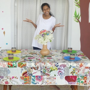 Peut inclure: Une nappe patchwork florale colorée recouvre une table dressée pour un repas. La table est ornée de quatre assiettes jaunes, vertes et bleues, chacune avec un verre décoré. Un vase de fleurs blanches est au centre.