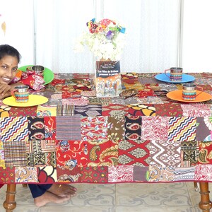 Peut inclure: Une nappe de patchwork color&eacute;e avec une base rouge et divers motifs dans des tons de brun, jaune, bleu et vert. Un livre intitul&eacute; "Les Filles de l'Etoile" est sur la table, ainsi que trois tasses et soucoupes color&eacute;es.