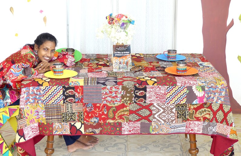 Peut inclure: Une nappe de patchwork color&eacute;e avec une base rouge et divers motifs dans des tons de brun, jaune, bleu et vert. Un livre intitul&eacute; "Les Filles de l'Etoile" est sur la table, ainsi que trois tasses et soucoupes color&eacute;es.