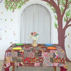 Peut inclure: Une nappe de table rouge en patchwork avec un centre de table floral color&eacute; sur une table avec trois assiettes. La table est dress&eacute;e contre un mur blanc avec un arbre peint et un rideau blanc.