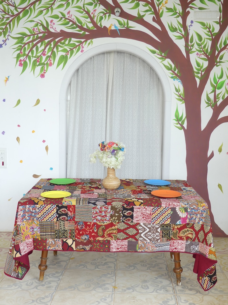 Peut inclure: Une nappe de table rouge en patchwork avec un centre de table floral color&eacute; sur une table avec trois assiettes. La table est dress&eacute;e contre un mur blanc avec un arbre peint et un rideau blanc.
