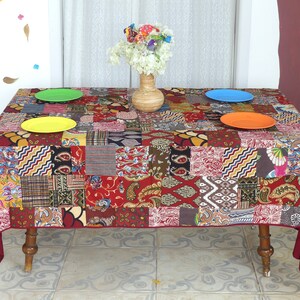 Peut inclure: Une nappe de table rouge en patchwork avec une vari&eacute;t&eacute; de motifs color&eacute;s. La nappe est pos&eacute;e sur une table avec quatre assiettes et un vase de fleurs.