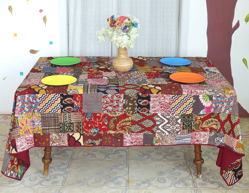Peut inclure: Une nappe de table rouge en patchwork avec une vari&eacute;t&eacute; de motifs color&eacute;s. La nappe est pos&eacute;e sur une table avec quatre assiettes et un vase de fleurs.
