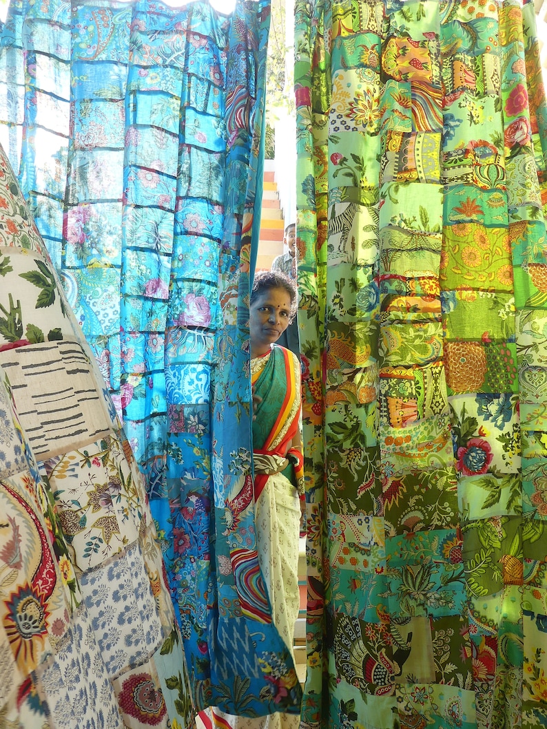 May include: A woman stands in front of a colorful patterned curtain. The curtain is made of many different fabrics sewn together. The woman is wearing a long, flowing dress with a floral pattern. She has dark hair and is looking at the camera.