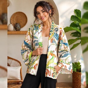 Peut inclure: Une femme porte une veste de style kimono crème avec un motif coloré et complexe. La veste présente des motifs bleus, verts, jaunes et rouges. Elle porte également un haut blanc et un pantalon noir. La veste a des manches larges.