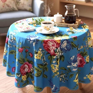 Peut inclure: Une table ronde recouverte d'une nappe bleue à motifs floraux. Une tasse de café, une soucoupe et un petit carnet sont sur la table. Un plateau avec une cruche de liquide et un sucrier est posé sur la table. Une chaise est en arrière-plan.