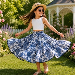 Peut inclure: Une jeune personne porte un haut court blanc, une jupe à motifs floraux bleus et blancs, des lunettes de soleil et un chapeau de paille. La jupe a un design fluide. Le fond présente un jardin avec des fleurs et un gazebo blanc.