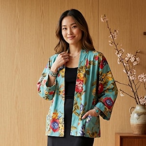 Puede incluir: Un kimono floral turquesa con un vibrante estampado de flores rojas, amarillas y rosas. El kimono tiene mangas largas y bolsillos. La mujer lleva un top y una falda negros.