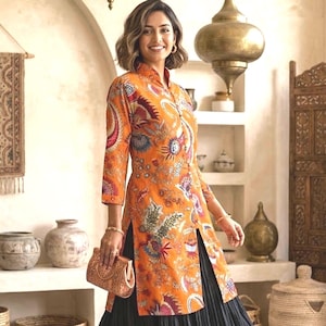 May include: A long-sleeved orange tunic featuring a vibrant, multi-colored floral and paisley print with a high collar and a front slit, paired with a black pleated skirt and a small, textured tan clutch bag held in the hand.