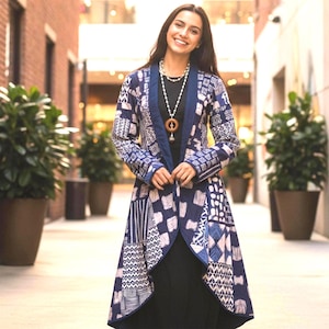 Peut inclure: Une femme porte une longue veste ouverte sur le devant avec un motif patchwork bleu marine et blanc. La veste a un col châle et des manches longues. Elle porte également un haut noir et un collier avec un pendentif en bois.