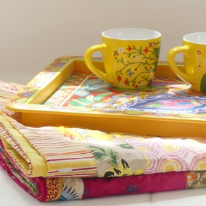 K&ouml;nnte beinhalten: Zwei gelbe Keramikbecher mit Blumendesigns stehen auf einem gelben Tablett mit einem bunten Muster. Das Tablett liegt auf einem Stapel bunter Stoffquadrate.