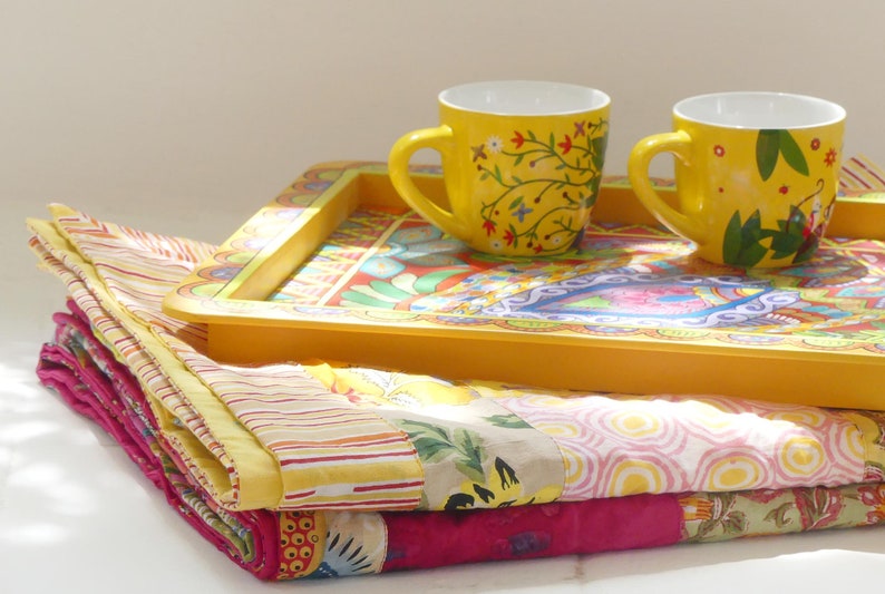 K&ouml;nnte beinhalten: Zwei gelbe Keramikbecher mit Blumendesigns stehen auf einem gelben Tablett mit einem bunten Muster. Das Tablett liegt auf einem Stapel bunter Stoffquadrate.