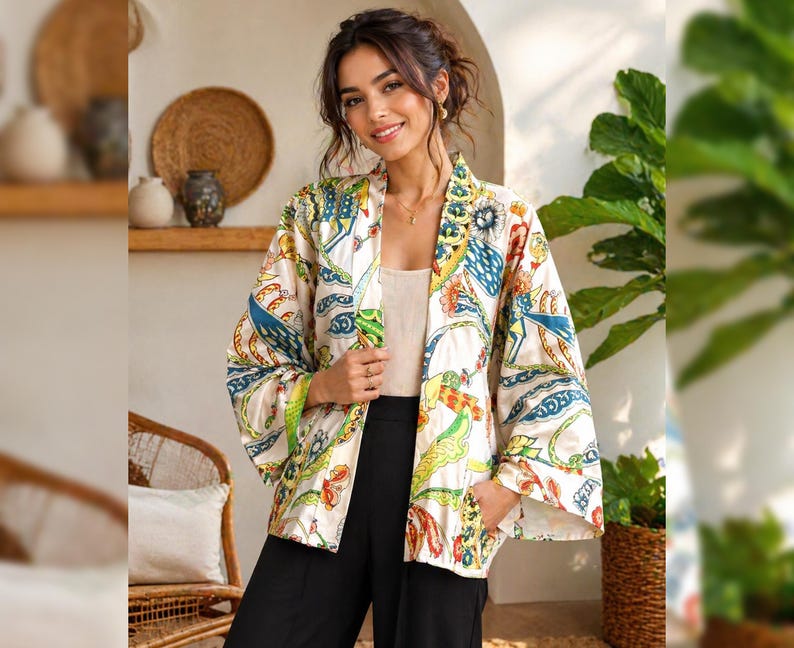 K&ouml;nnte beinhalten: Eine cremefarbene Kimono-Jacke mit einem farbenfrohen, gemusterten Design mit blauen, gelben und roten Akzenten. Die Jacke hat weite &Auml;rmel und ist vorne offen. Das Model tr&auml;gt eine schwarze Hose und ein cremefarbenes Oberteil.