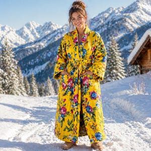 May include: A long, yellow floral robe with a tie waist and pockets. The robe features a vibrant pattern of pink, blue, and green flowers. The robe is worn outdoors in a snowy, mountainous landscape.