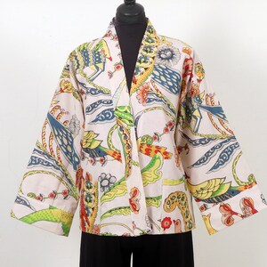 K&ouml;nnte beinhalten: Eine cremefarbene Jacke im Kimono-Stil mit einem lebendigen, farbenfrohen Druck. Das Muster zeigt V&ouml;gel, Blumen und abstrakte Formen in Blau-, Gelb-, Gr&uuml;n- und Rott&ouml;nen. Die Jacke hat weite &Auml;rmel und eine lockere Passform.