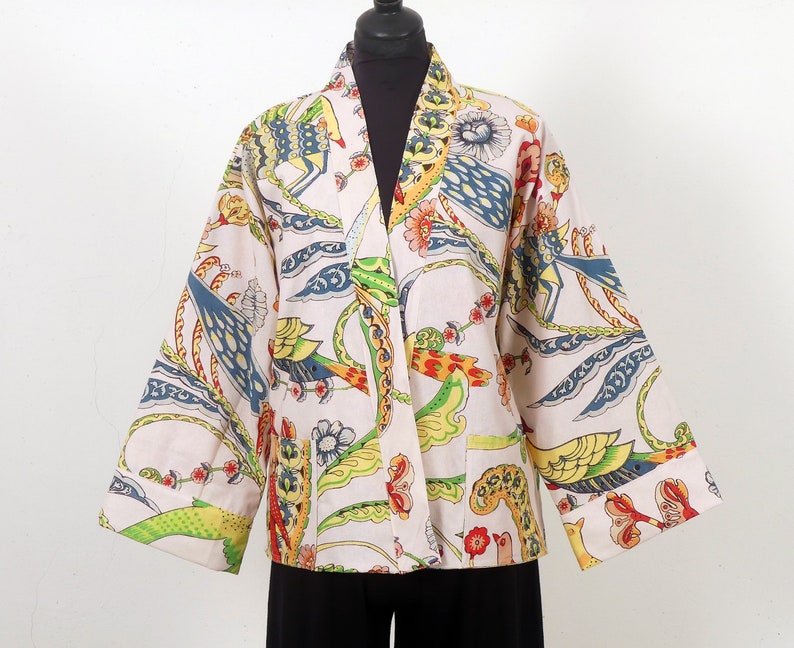 K&ouml;nnte beinhalten: Eine cremefarbene Jacke im Kimono-Stil mit einem lebendigen, farbenfrohen Druck. Das Muster zeigt V&ouml;gel, Blumen und abstrakte Formen in Blau-, Gelb-, Gr&uuml;n- und Rott&ouml;nen. Die Jacke hat weite &Auml;rmel und eine lockere Passform.