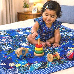 Peut inclure: Une courtepointe à motif floral bleu avec un tout-petit jouant avec un jouet à empiler en bois, un éléphant en bois à roues bleues, un hochet roulant en bois et une balle en tissu colorée. La courtepointe a une bordure rayée bleue et blanche.