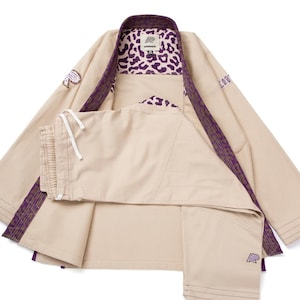 Op de afbeelding: Een beige vechtsport-gi met een paarse luipaardprint voering op de kraag en aan de binnenkant. Het jasje heeft lange mouwen met drievoudig gestikte boorden, en de bijpassende broek heeft een witte taille met trekkoord en een klein paars logo.