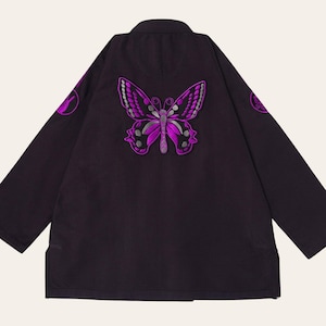 Puede incluir: Una chaqueta negra de manga larga con un gran bordado de mariposa en color morado y plateado centrado en la espalda. Las mangas muestran parches circulares bordados en morado, y la prenda tiene un dobladillo recto con aberturas laterales.