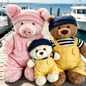 Puede incluir: Tres animales de peluche: un cerdo rosa grande con gorro y peto rosa sobre una camiseta a rayas, un oso pardo grande con gorro azul marino y peto amarillo sobre una camiseta a rayas, y un oso blanco pequeño con gorro azul marino y peto amarillo.