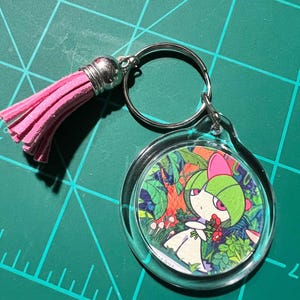 May include: A circular clear acrylic keychain featuring a colorful illustration of a green and white character with pink accents holding red flowers in a forest setting, attached to a silver metal ring with a soft pink faux suede tassel.