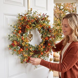 May include: A circular autumn wreath featuring green eucalyptus leaves, small orange pumpkins, dark brown pinecones, and clusters of red and yellow berries. The wreath is mounted on a white door with a gold-colored metal door knocker in the center.