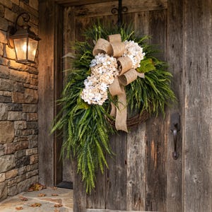 May include: A teardrop-shaped wreath featuring a base of woven brown twigs, adorned with lush green fern-like foliage, clusters of cream-colored hydrangea blossoms, and a large, decorative tan burlap ribbon tied in a multi-loop bow at the top.