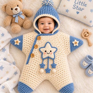Op de afbeelding: Een crème en blauwe gehaakte stervormige babyslaapzak met capuchon, voorzien van een lachende sterapplicatie, drie houten knopen en blauwe pomponaccenten. Het kledingstuk wordt omringd door een teddybeer, een kussen met de tekst Little Star en babyslofjes.