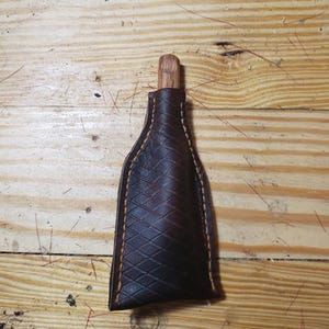 Könnte beinhalten: Eine dunkelbraune Lederscheide mit rautenförmiger Textur und dicken beigen Nähten an den Kanten. Ein kleiner, heller Holzgriff ragt aus der oberen Öffnung des Lederetuis heraus, das passgenau für den darin befindlichen Gegenstand geformt ist.