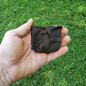 Peut inclure: Une petite pochette en cuir marron foncé avec une surface texturée et patinée. La pochette présente une fermeture à rabat simple sécurisée par une fine lanière en cuir qui s'enroule sur le devant, offrant un design compact pour petits objets.