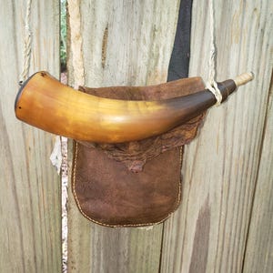 Puede incluir: Un cuerno de pólvora curvo de color marrón claro a oscuro con una punta clara, unido a una bolsa de cuero marrón oscuro. El conjunto cuenta con una correa de cordón trenzado de color claro para llevarlo, con costuras visibles en los bordes.