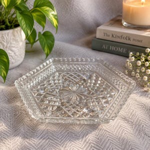 May include: A clear, hexagonal glass tray featuring a pressed diamond pattern and a central starburst design. The tray has a raised, scalloped edge with a beaded texture, reflecting light across its faceted surface to create a decorative display.