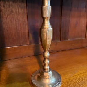 May include: A polished brass candlestick holder featuring a wide circular base, a slender central stem with decorative turned detailing, and a flared top cup designed to securely hold a single taper candle in an upright position.