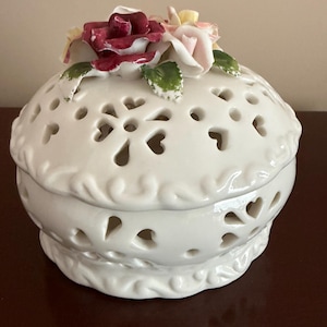 May include: A round white ceramic trinket box with a removable lid featuring a cut-out heart pattern. The top of the lid is decorated with sculpted porcelain roses in shades of deep red, pale pink, and yellow, accented with green leaves.