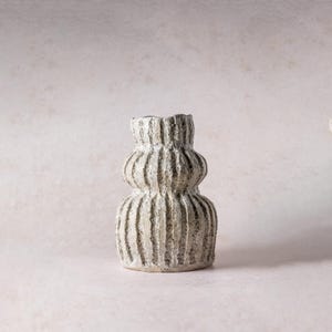 Puede incluir: Un pequeño jarrón de cerámica de color blanco roto con una superficie texturizada y moteada, y un distintivo patrón de nervaduras verticales. El jarrón presenta una silueta redondeada y escalonada con un cuello estrecho y una base bulbosa.