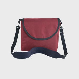 May include: A maroon messenger bag featuring a rounded flap closure with dark navy blue trim. The bag includes a wide, adjustable dark navy blue shoulder strap attached with black plastic swivel clips on both sides, providing a functional design.
