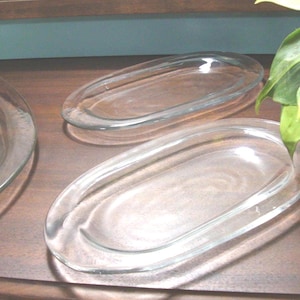 May include: Two clear glass oval serving platters with smooth, rounded edges and a shallow basin. The dishes are transparent, featuring a simple, unadorned design with a slightly raised rim, reflecting light across their polished surfaces.