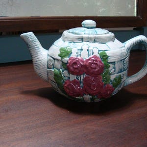 Könnte beinhalten: Eine Keramik-Teekanne mit einem hellblauen und weißen Korbgeflechtmuster und einem weißen Lattenzaun-Design. Die Teekanne ist auf der Vorderseite mit dreidimensionalen dunkelrosa Rosen und grünen Blättern sowie einem passenden Deckel verziert.
