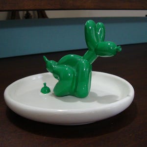 May include: A glossy green ceramic balloon dog figurine is attached to the center of a round white ceramic tray. The dog is sculpted in a seated position with a small green drop shape placed on the tray surface near the dog's hindquarters.