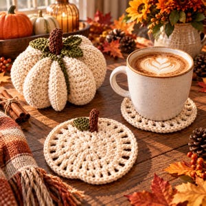 May include: A cream-colored crocheted pumpkin with a brown stem and green leaves, a matching flat crocheted pumpkin coaster, and a speckled ceramic mug filled with coffee featuring latte art, all arranged on a wooden surface with autumn decor.
