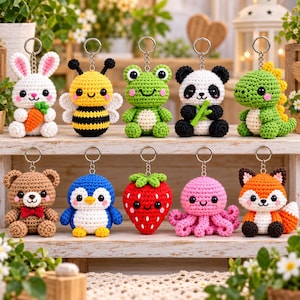Può includere: Una collezione di dieci portachiavi all'uncinetto a forma di animali e frutti, tra cui un coniglio bianco con carota, un'ape gialla e nera, una rana verde, un panda con bambù, un dinosauro verde, un orso marrone, un pinguino blu, una fragola rossa, un polpo rosa e una volpe.