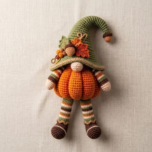 May include: A crochet gnome doll featuring a large orange pumpkin-shaped body, a tall olive green pointed hat, and striped legs. The hat is decorated with a small brown acorn, orange berries, and a green leaf. The doll has a round beige nose.